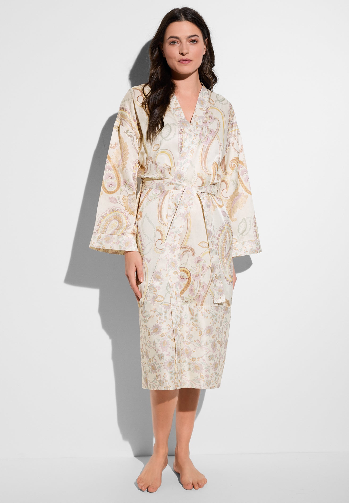 Woman standing in a cream paisley robe, facing the camera against a white background, bare feet visible.