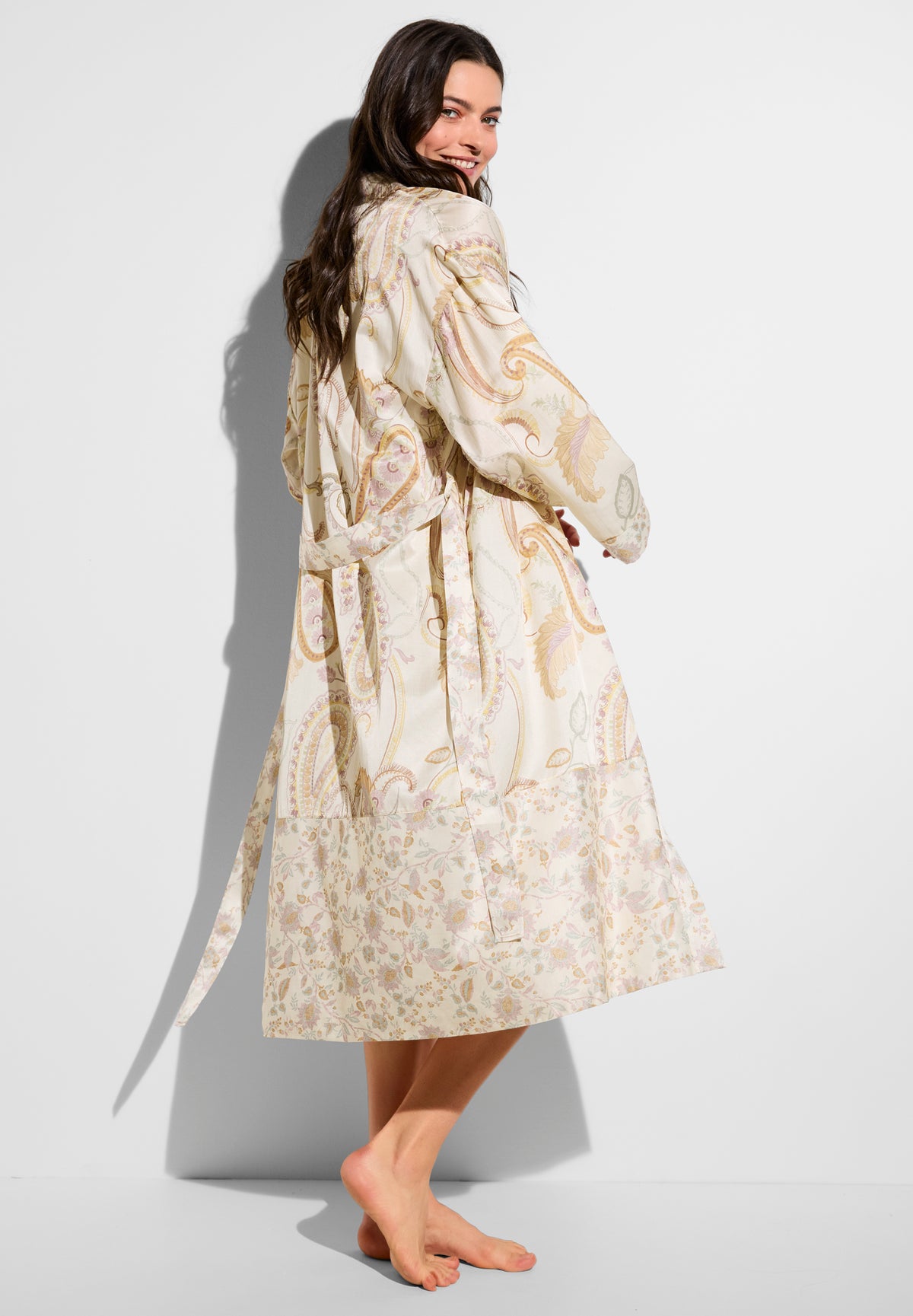 Woman in a cream paisley robe, shown from the back/side, smiling with long dark hair, bare feet against a white background.