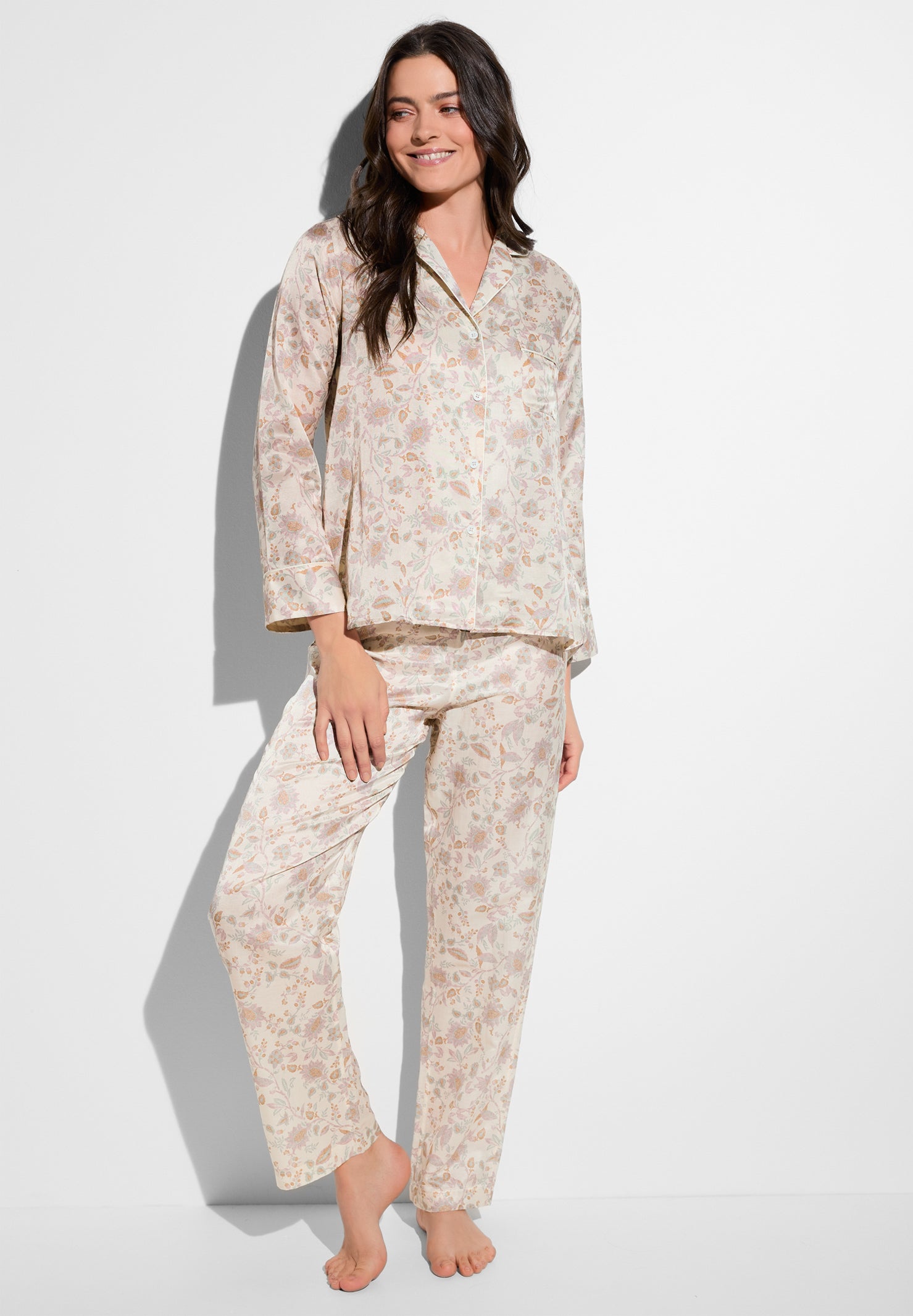 Smiling woman wearing pale pink floral pajama set, standing barefoot against a white background.