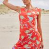 Woman on a sandy beach wearing a bright red floral dress, her hair pulled back, adjusting a ponytail with one hand.