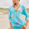 Woman in a bright turquoise, printed beach dress posing on a sandy beach with hands on her hips.