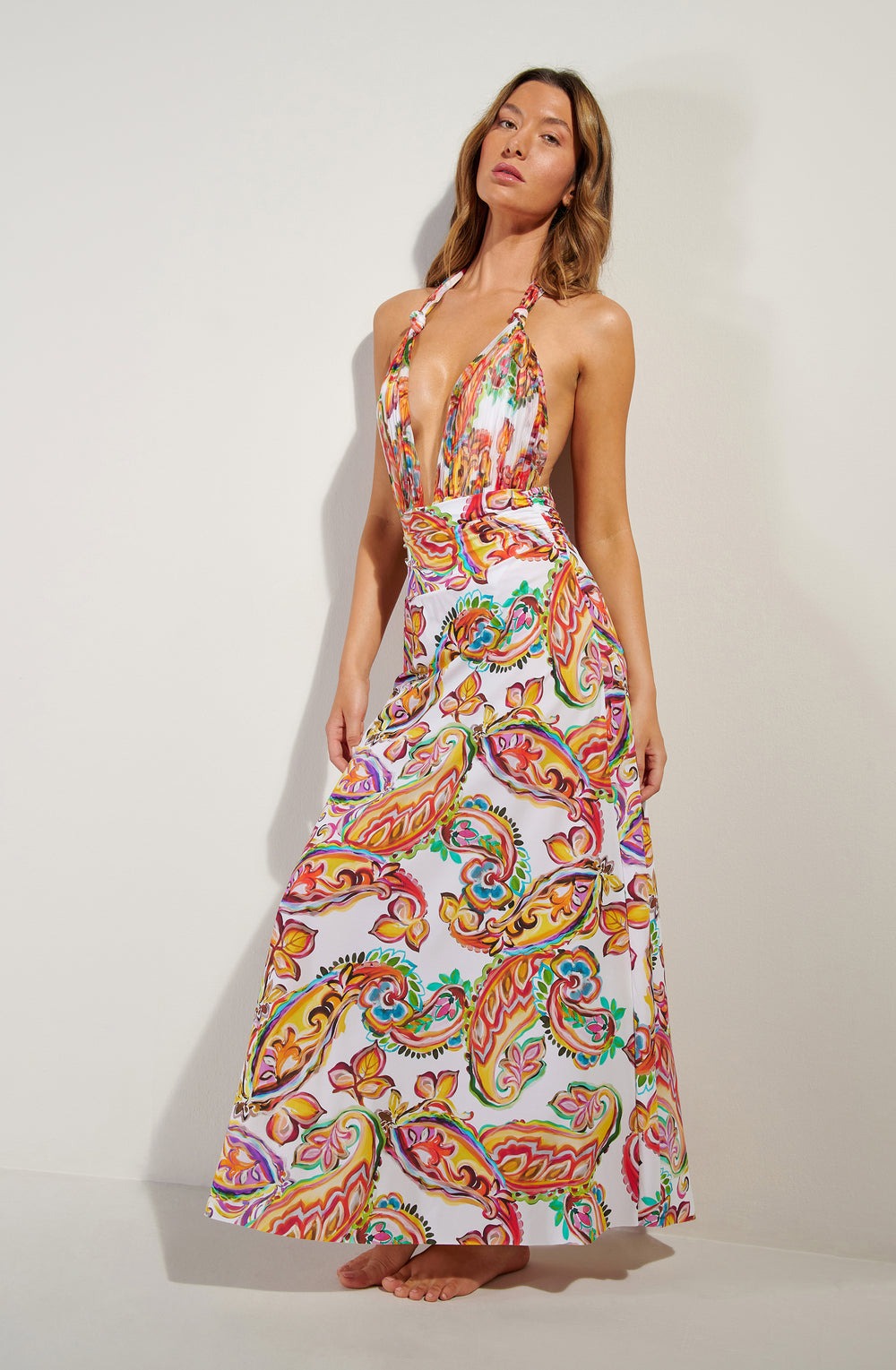 Model wearing a vibrant paisley-print halter maxi dress with a plunging V-neck, floor-length skirt, barefoot against a pale wall.