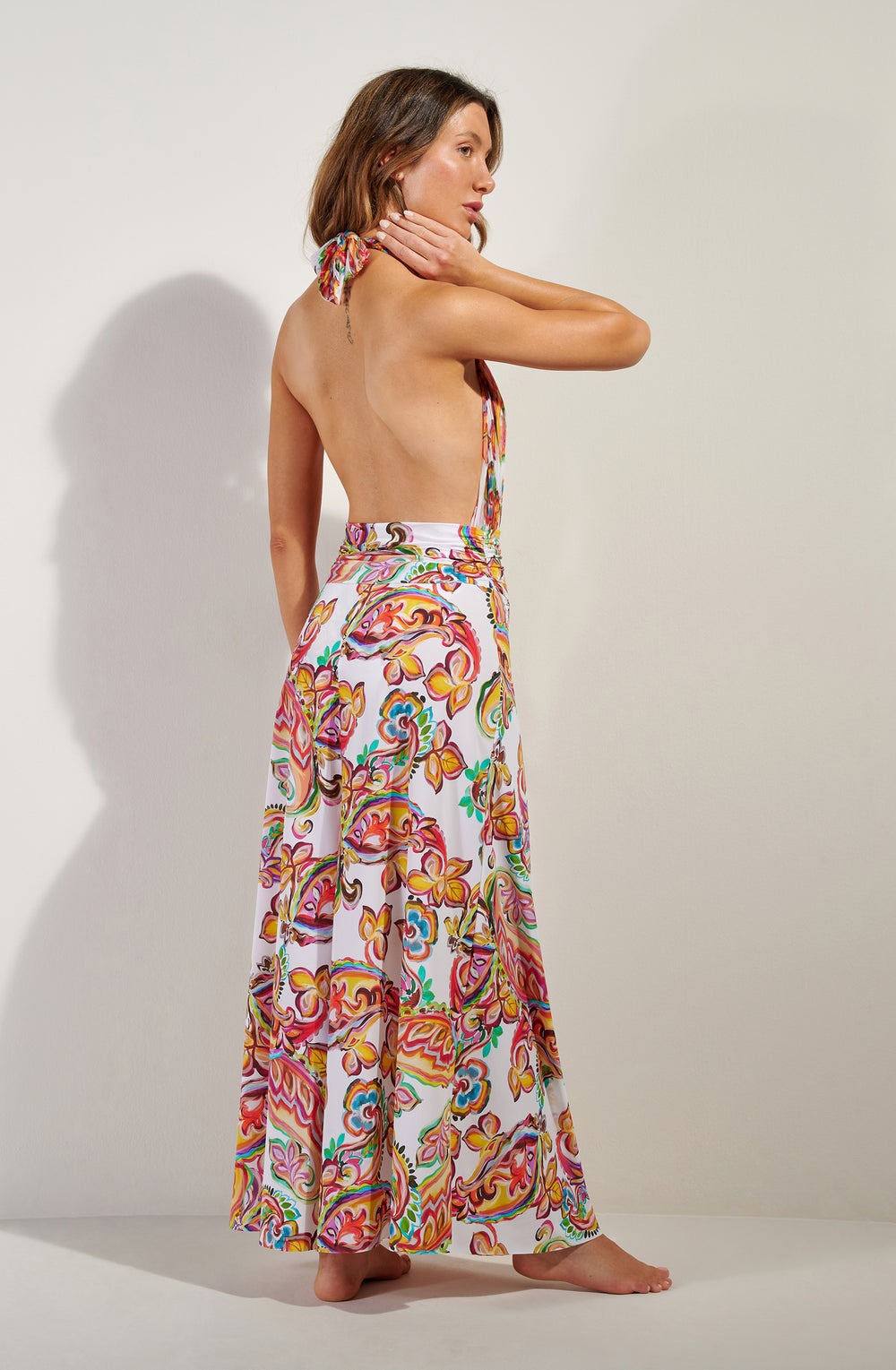 Model wearing a backless halter floral maxi dress, standing sideways with one hand at her neck, barefoot against a light background.