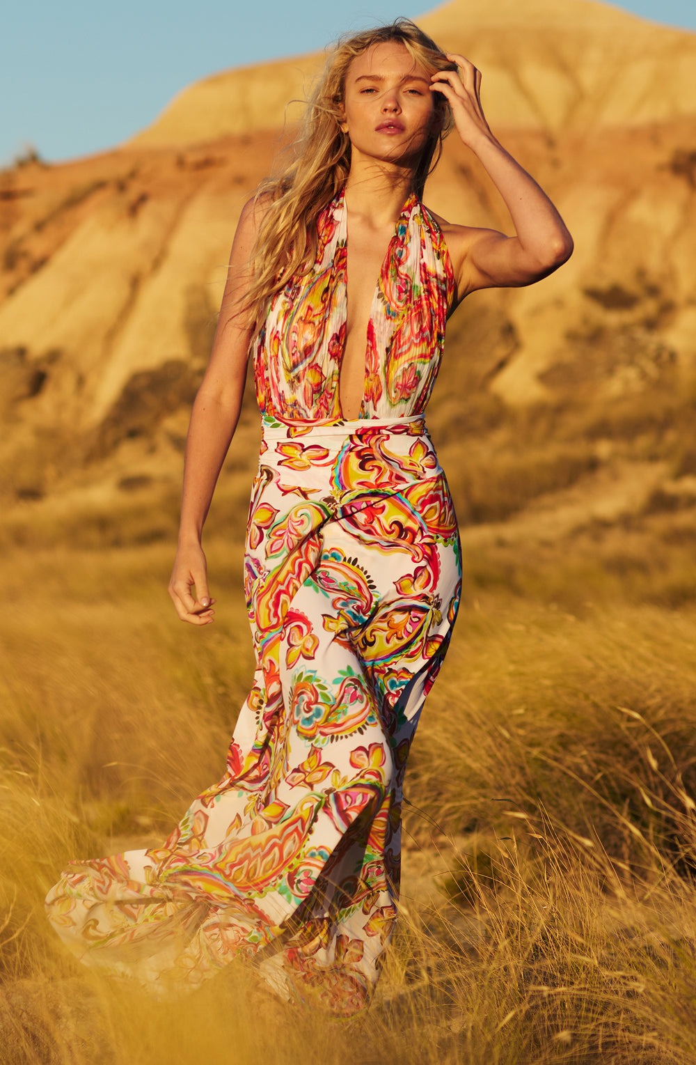 Woman with long blonde hair walking through a desert in a bright, paisley-patterned halter maxi dress in golden hour light.