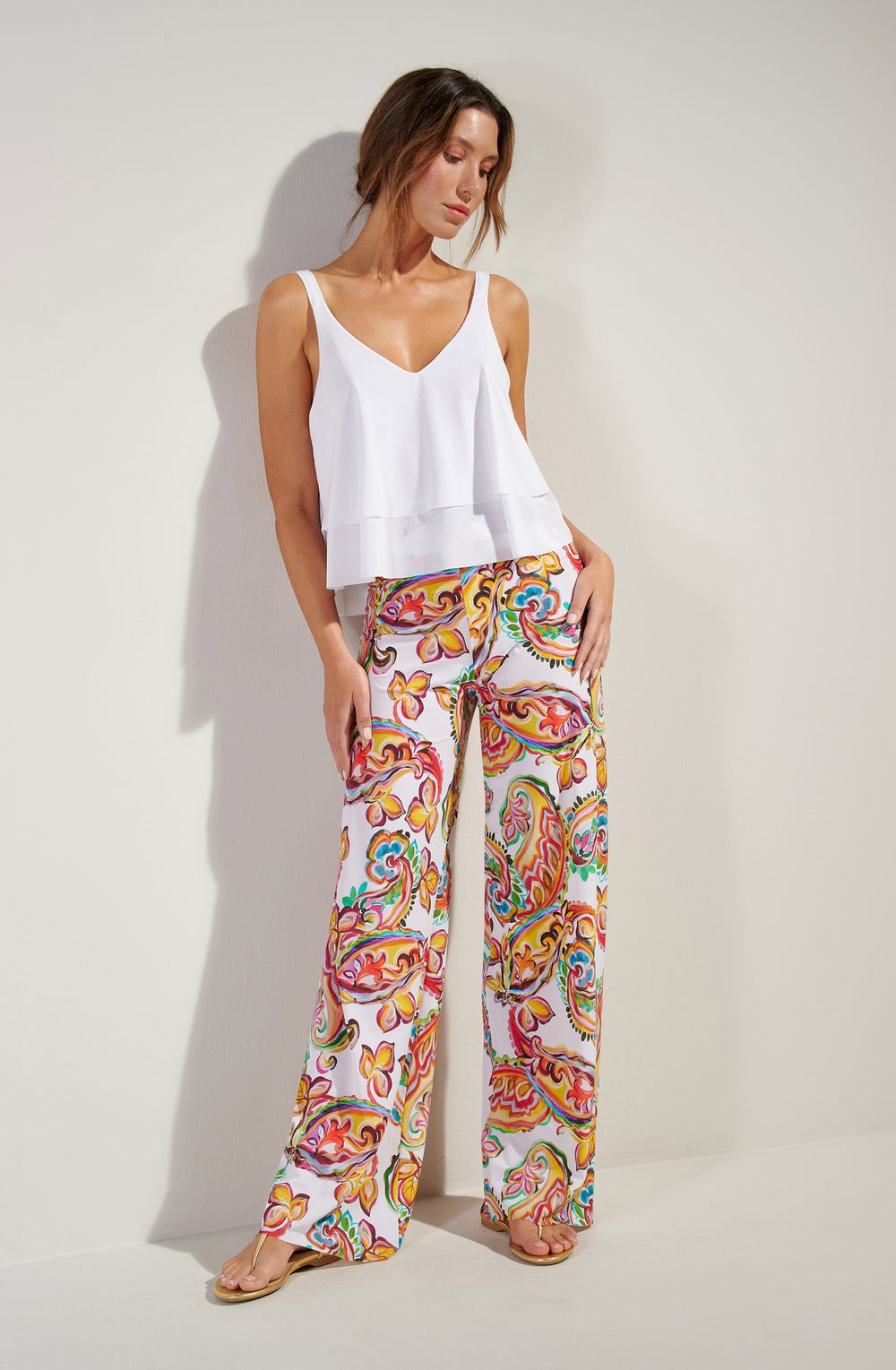 Woman wearing a white sleeveless layered top and bright multicolor paisley-patterned wide-leg pants, standing against a white wall.