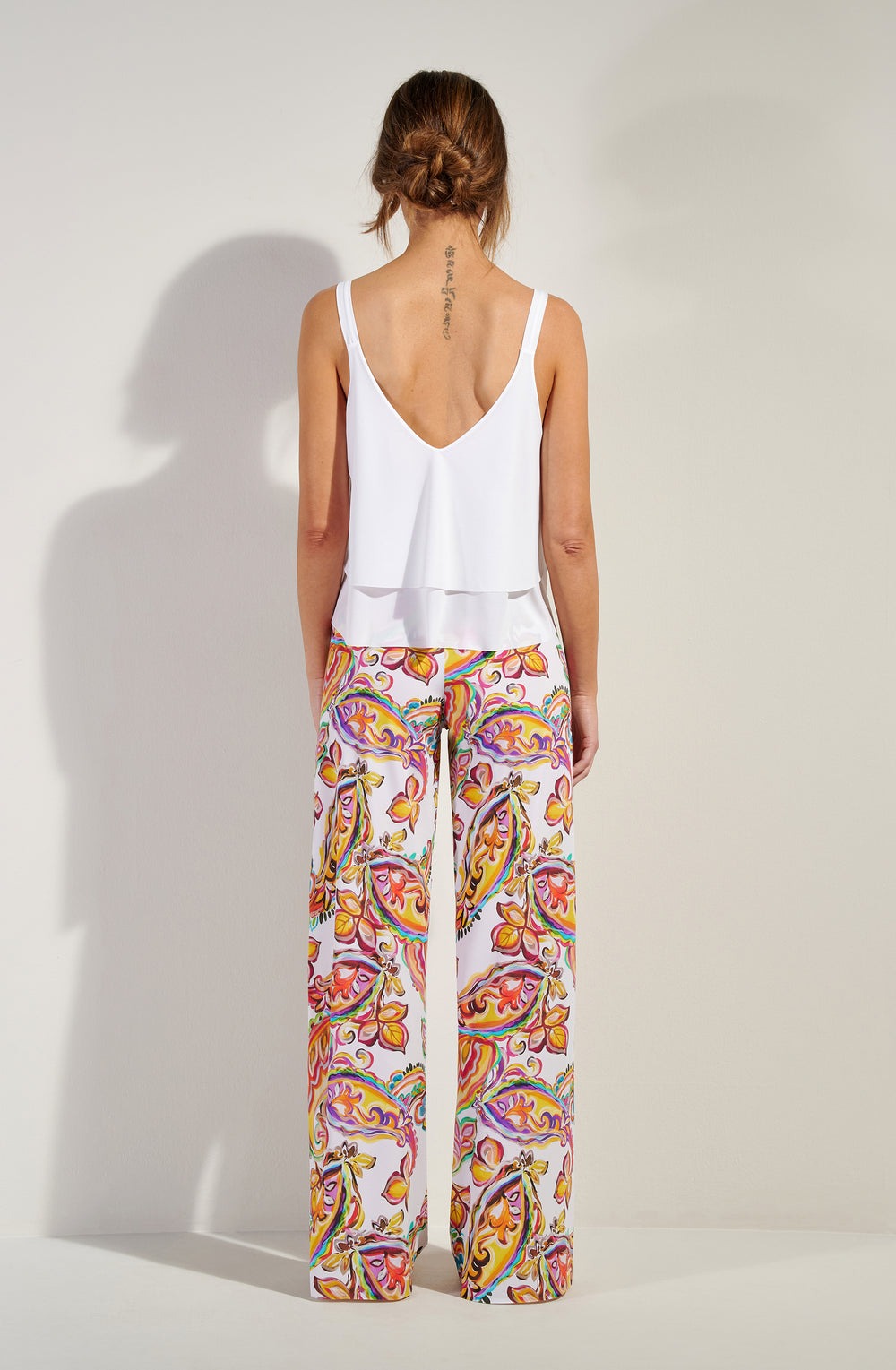 Back view of a woman in a white sleeveless top and vibrant paisley-patterned wide-leg pants, with a tattoo along her upper spine.