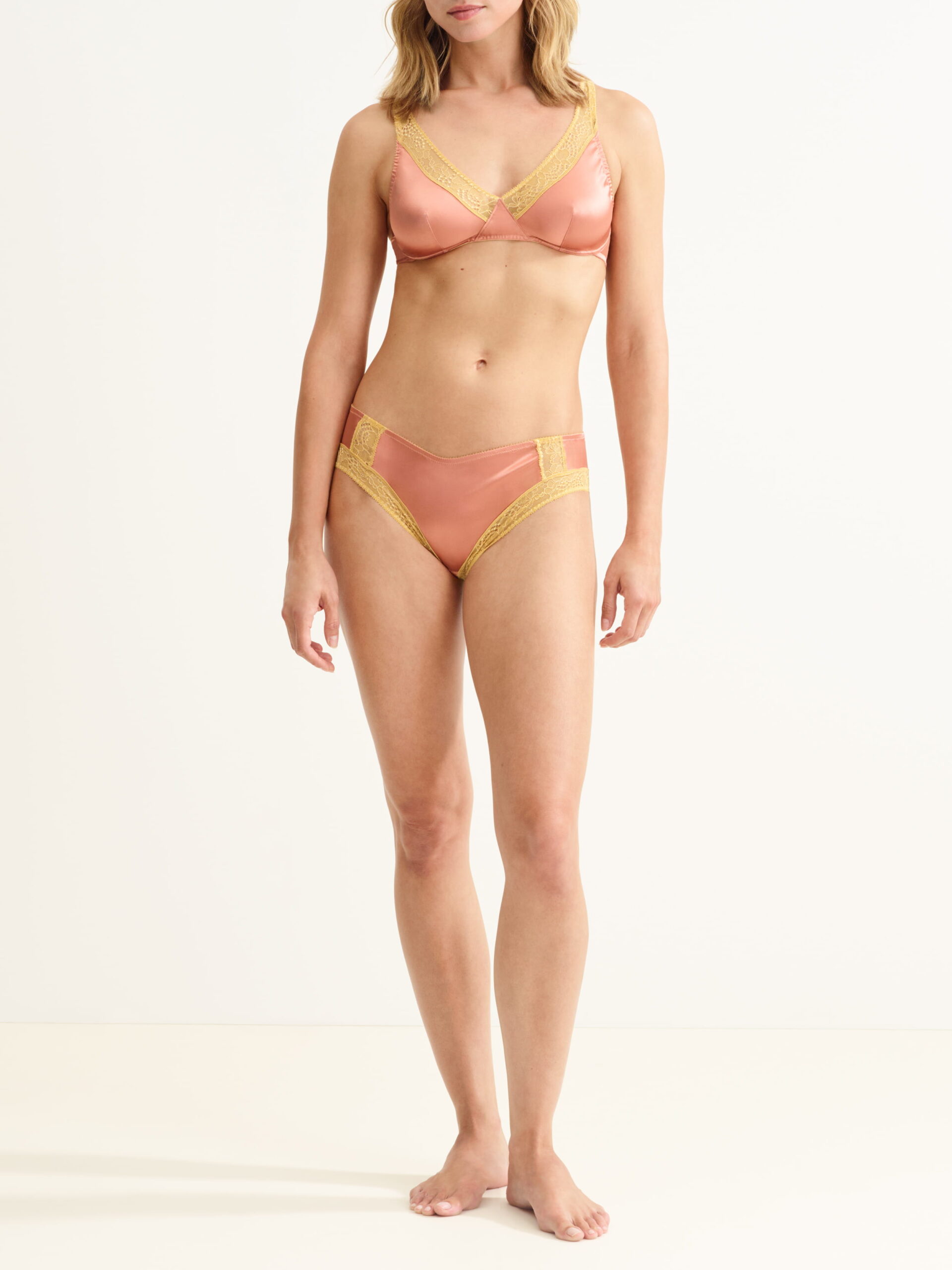 Woman wearing a pink satin bra and matching panties with gold lace trim, standing against a light background.