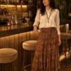 Woman leaning on a bar counter in a warmly lit bar, wearing a cream embroidered blouse and a long paisley skirt, with bottles in the background.