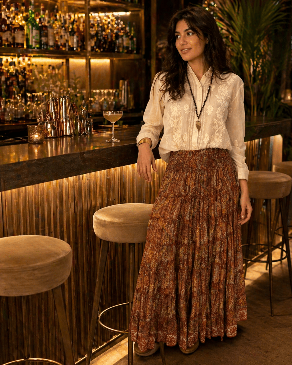Woman leaning on a bar counter in a warmly lit bar, wearing a cream embroidered blouse and a long paisley skirt, with bottles in the background.