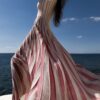 Woman in a pink and white striped dress stands on a rocky beach, the ocean and blue sky behind her.
