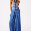 Model in a blue denim halter top with crochet panels and wide-leg jeans, wearing gold floral earrings, standing against a white studio backdrop.",