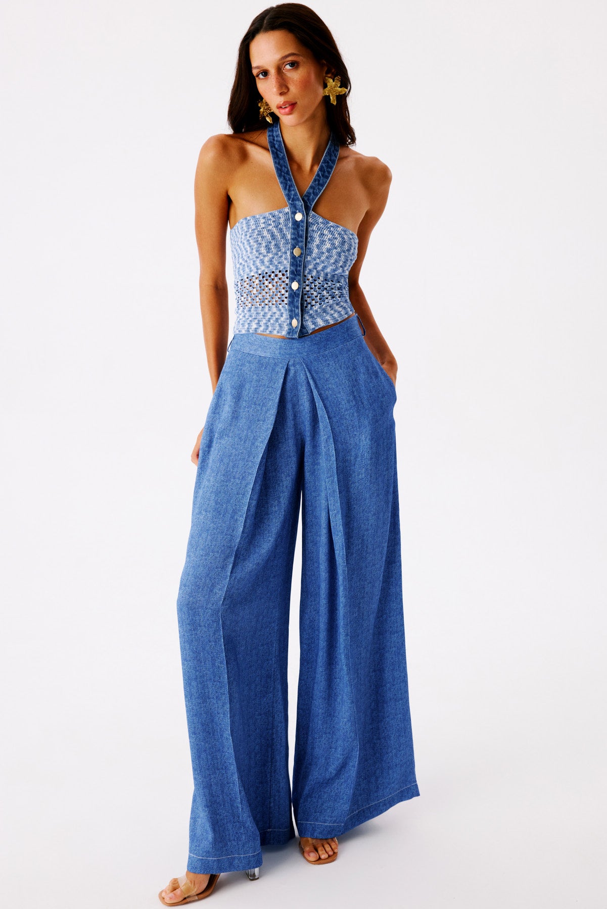 Model in a blue denim halter top with crochet panels and wide-leg jeans, wearing gold floral earrings, standing against a white studio backdrop.",