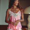 Woman wearing pink satin camisole and shorts with white lace trim, standing indoors in a softly lit room.