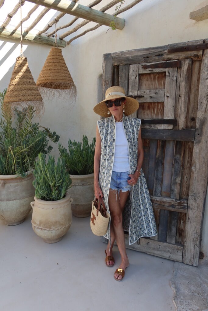 Woman in a wide-brim straw hat and sunglasses poses by a weathered wooden door, wearing a patterned sleeveless vest over a white top with denim shorts and brown sandals, carrying a woven tote.